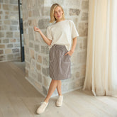 Woman wearing a perfect white tee and brown striped, sporty skirt standing in a room with stone walls and curtains.