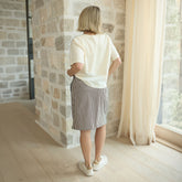 Back view of woman wearing knee-length skirt in brown stripes with an ivory tee and sneakers against a stone wall.