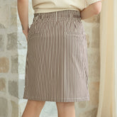 Back view of woman wearing a knee length, brown striped skirt with back pockets against a blurred stone wall background
