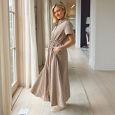 Woman in a long, brown gingham dress standing in a well-lit room with large windows.