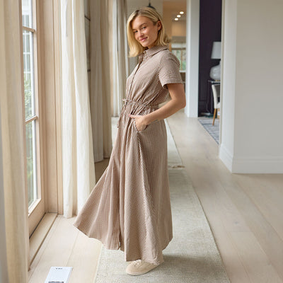 Woman in a long, brown gingham dress standing in a well-lit room with large windows.