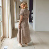 Woman in a brown gingham maxi dress standing in a bright room with large windows.
