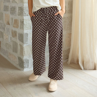 Woman wearing brown polka dot, wide leg pants with sneakers standing against a stone wall.