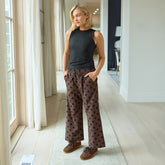 Woman wearing a black sleeveless top and patterned brown women's pants standing in a bright room with large windows.