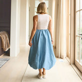 Woman wearing a white tank top and blue midi skirt standing in a bright room with large windows.