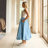 Woman wearing a light chambray skirt and white top standing in a bright room with large windows.
