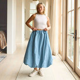 Woman wearing a cropped, white tank top and blue bubble skirt standing in a well-lit room with large windows.