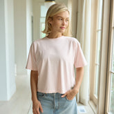 Woman wearing a light pink t-shirt and denim skirt standing in a bright room with large windows.