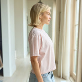 Side view of a woman wearing a boxy women's t-shirt in baby pink with a denim skirt facing large windows.