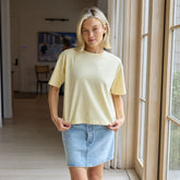 Women’s butter yellow t-shirt, The Perfect Tee, worn with a denim skirt in a bright indoor setting with large windows.