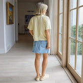 Woman standing by a large window in a bright room, wearing a boxy t-shirt in butter yellow and denim skirt.