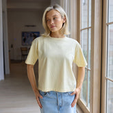 Woman standing in a large room wearing a beautiful, light yellow t-shirt and denim skirt.