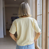 Back view of woman wearing a perfectly cropped, light yellow t-shirt and denim skirt standing in a home.