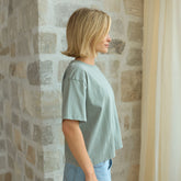 Side view of woman wearing The Perfect Tee, sage green t-shirt standing against a stone wall.