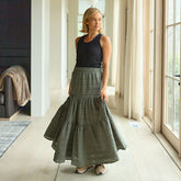 Woman wearing a long green skirt and black top in a bright room with large windows.