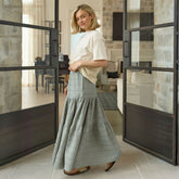 Woman in a white top and taupe skirt standing in a modern interior setting with large glass doors.