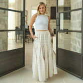 Woman in a white top and off white maxi skirt standing in a modern interior setting.
