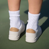Back view of Sorel Ona Drift sneakers in Sea Salt and Honest Beige with white ribbed socks on a green background
