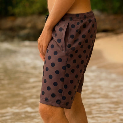 Side view of man wearing brown swim shorts for men with black polka dots on a blurred ocean background