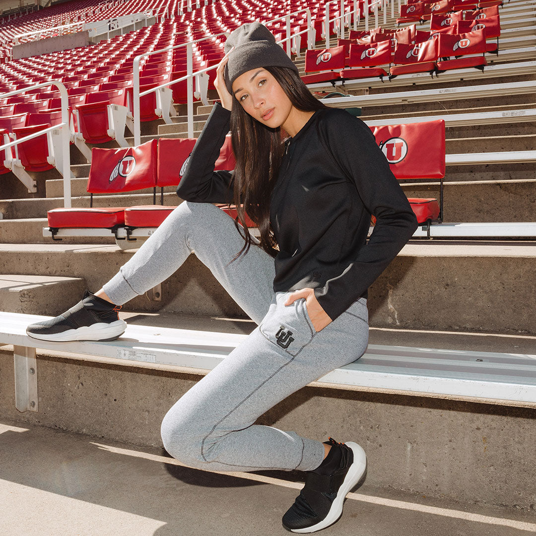 Woman sitting on stadium seats wearing grey sweatpants with an embroidered U of U logo, paired with a black top and black hat.