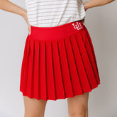 A person wearing a red pleated tennis skirt with a built-in shorts and a high-waisted fit. The skirt features an embroidered U of U logo.