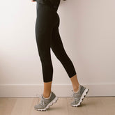 Side view of black Intention Flow Capris showcasing the high-waisted capri length on a model with gray sneakers indoors