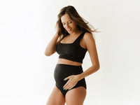 Front view of a black maternity two-piece swimsuit on a pregnant model, showcasing its bump-friendly fit against a white background