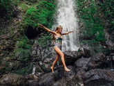 Woman poses in a green patterned bikini top and striped high-waisted bottoms on rocks before an Oahu waterfall