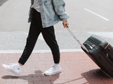 Person walking with a black rolling suitcase on a city sidewalk, wearing black pants, a denim jacket, and white sneakers