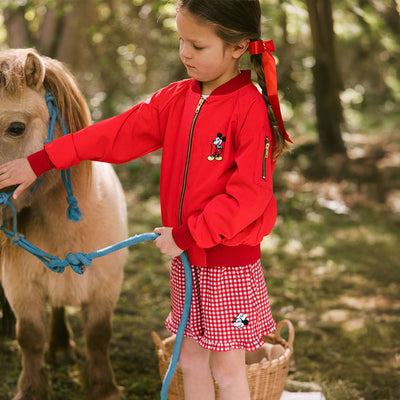 Young girl petting a small pony and wearing a red Disney jacket for kids with Mickey Mouse embroidery and a red gingham Disney shorts for kids. 