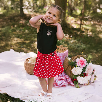 Young girl in a red polka dot skirt Minnie Mouse skirt and black Minnie Mouse top for kids standing on a picnic blanket with flowers and baskets in a park.