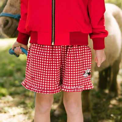 Close up of child wearing red gingham Minnie Mouse shorts for kids with a blurred pony in the background.