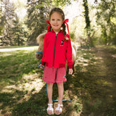 Young girl standing in front of a pony wearing a red kid's Mickey jacket and red gingham kid's Disney shorts with Minnie Mouse embroidery. 