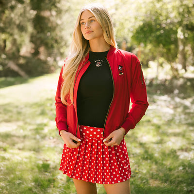 Woman standing in a park wearing a Minnie Mouse tank top, red Mickey Mouse bomber jacket, and Minnie Mouse tennis skirt in red polka dots.