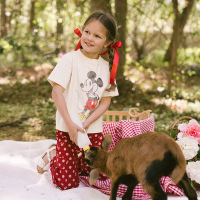 Young girl on a picnic blanket feeding a baby goat and wearing a cute Mickey Mouse tee and Disney pants for kids. 