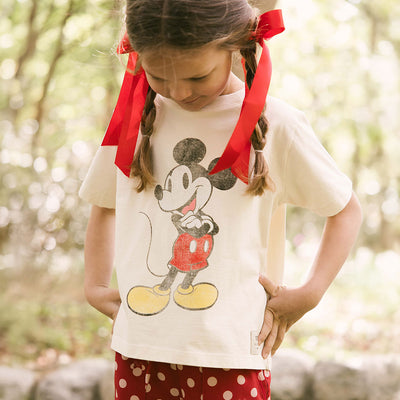 Young girl wearing a kids Mickey t-shirt and kids Disney pants with red bows in her hair on a blurred natural background