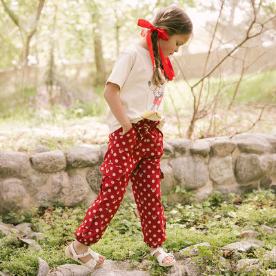 Young girl standing in a garden wearing a Mickey Mouse t shirt and red polka dot kid's Disneyland pants with lots of pockets. 