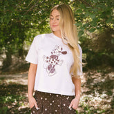 Close up of woman wearing a white Minnie Mouse tee with neutral Disney shorts on a blurred greenery background.