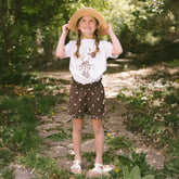 Young girl standing in a forest wearing a cute Disney kids outfit—kids Minnie Mouse tee with kids Minnie Mouse shorts in brown. 