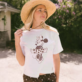 Woman wearing a flattering, neutral women's Disney shirt with Minnie Mouse graphic against a blurred greenery background.