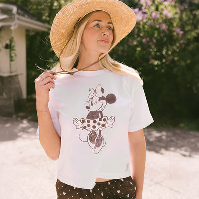 Woman wearing a flattering, neutral women's Disney shirt with Minnie Mouse graphic against a blurred greenery background.