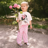 Child wearing a Minnie Mouse shirt and pink checkered pants, holding flowers outdoors.