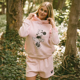 Woman standing in a forest wearing the perfect Disneyland outfit—women's Minnie sweatshirt and women's Disney shorts in light pink.