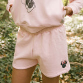 Close up of woman wearing pink women's Disney shorts with embroidered Minnie Mouse icon on a blurred greenery background.