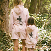 Mom and daughter wearing matching Disney outfits—pink Minnie Mouse sweatshirts and pink Minnie Mouse shorts in a forest setting.