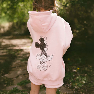Back view of child wearing pink Disneyland sweatshirt with Minnie Mouse on the back on a blurred green background.