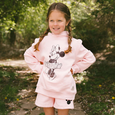 Young girl wearing a pink Minnie Mouse hoodie and shorts with a Minnie Mouse design, standing outdoors.