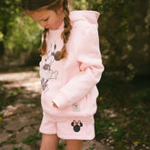 Child wearing a pink hoodie with Minnie Mouse design and pink shorts with Minnie embroidery on a blurred greenery background. 