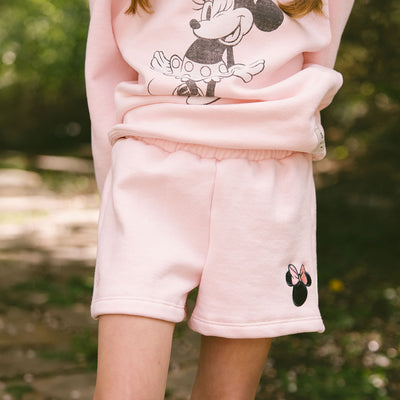 Close up of child wearing cute kid's Disney outfit—pink Minnie Mouse shorts and kid's pink Minnie Mouse hoodie on a blurred green background. 
