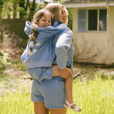 Woman holding a child on her back in a green field wearing matching Disney outfits—blue kids Disney sweatshirt and matching shorts. 